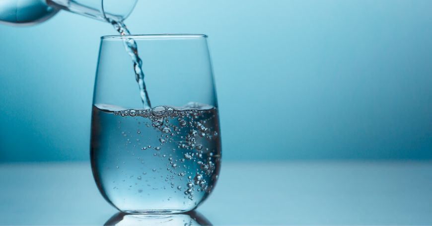 Un bicchiere d'acqua con bollicine, riempito da una caraffa, su uno sfondo blu. L'acqua frizzante può avere effetti negativi per chi soffre di colite, causando gnfiore addominale